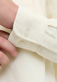 Baltas kordūrinių marškinių rankogalis su dviem apvaliais mygtukais ir tekstūruota raya; ranka reguliuojanti rankogalį.