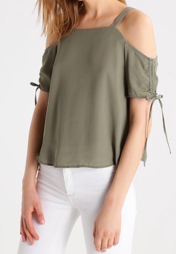 Olivově zelený top s krátkým rukávem, s výřezy na ramenou, zavázanými rukávy a volným střihem. Hladká textury a rovný lem, v kombinaci s bílými kalhotami.