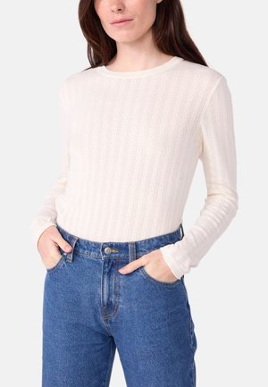 Femme portant un pull texturé blanc à manches longues et un jean bleu taille haute, les mains dans les poches, sur un fond uni.