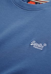 Blauwe katoenen t-shirt met een ronde hals en geborduurd wit logo op de linkerborst, met een zachte textuur en een casual ontwerp.