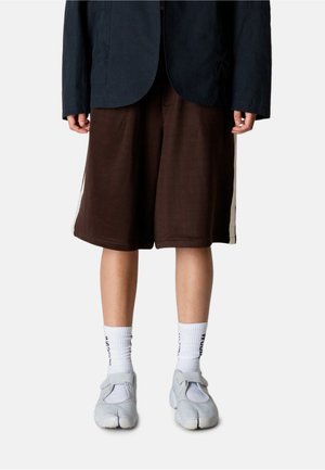 Person iført brune knælange shorts med vide ben, hvide sokker med sort tekst og grå sko med delt tå, stående mod en ensfarvet baggrund.