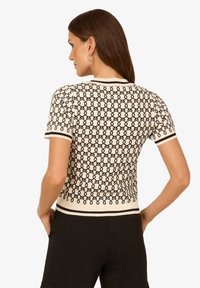 Leichtes Strickoberteil in Creme mit einem schwarzen geometrischen Muster, kurzen Ärmeln und einer figurbetonten Passform. Mit gerippten Details an den Bündchen und dem Saum.