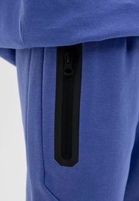 Pantaloni sportivi viola con una tasca zip nera sul lato, realizzati in tessuto morbido, caratterizzati da una texture liscia e un design affusolato.