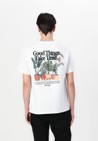 Camiseta de algodón blanca con un estampado gráfico de macetas y el texto "Las cosas buenas llevan tiempo" en la parte posterior. Mangas cortas, cuello redondo.