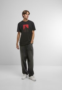 Mladý muž stojí s jednou rukou vo vrecku, oblečený v čiernom grafickom tričku, voľných tmavých džínsoch a bielych teniskách na jednoduchej pozadí.