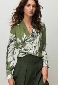 Žalia gėlėta palaidinė ilgomis rankovėmis ir V formos iškirpte, derinama su aukštakūnėmis žaliomis klostuotomis kelnėmis. Audinys pasižymi ryškiais juodais ir baltais gėlių raštais.