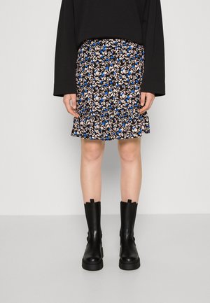 Persoon draagt een zwart shirt met lange mouwen, een rok met bloemenpatroon in blauw en beige, en zwarte laarzen tot halverwege de kuit, staand op een grijze vloer.