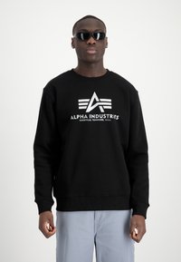Fekete pulóver kerek nyakkivágással, hosszú ujjal, elöl egy fehér grafikai logóval, amely az "ALPHA INDUSTRIES" feliratot és három elegáns vonalat ábrázol.