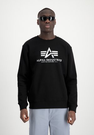 Svart hettegenser med rund hals, lange ermer, prydet med et hvitt grafisk logo "ALPHA INDUSTRIES" og tre stilige linjer på forsiden.
