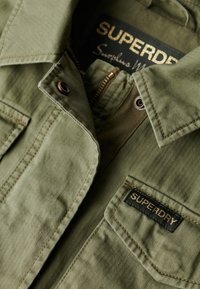 Olijf groene jas van textuurstof, voorzien van een ritsluiting en een patch met het "SUPERDRY" label. Heeft een kraag en knoopdetails.