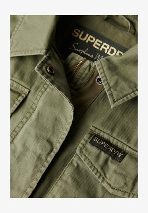 Giacca verde oliva realizzata in tessuto testurizzato, dotata di chiusura con zip e di una toppa con etichetta "SUPERDRY". Presenta un colletto e dettagli con bottoni.