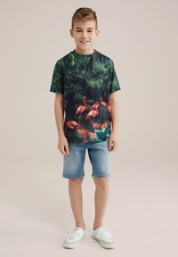 T-shirt med tropiskt tryck med gröna bladverk och rosa flamingor, parat med denimshorts och vita sneakers.