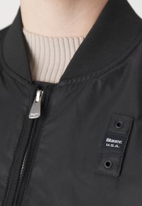 Černá bomber bunda s žebrovaným límcem, zapínáním na zip a štítkovou nášivkou. Hladká textilie s elegantním, minimalistickým designem.