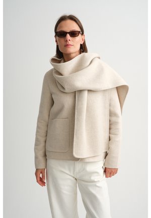 Femme portant une veste en laine crème, une grande écharpe assortie drapée sur l'épaule, un pantalon blanc et des lunettes de soleil fines et sombres, sur un fond uni.