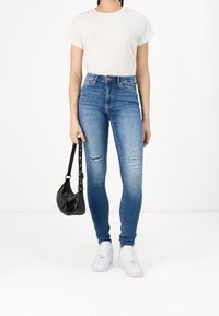 Jeans ajustados de tiro alto en azul con ligeros desgastes, combinados con una camiseta blanca y zapatillas blancas. Sosteniendo un bolso negro.