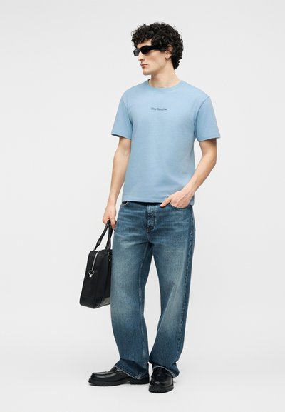 Άνδρας που φορά μπλε μπλούζα, φαρδιά μπλε τζιν, μαύρα παπούτσια, μαύρα γυαλιά ηλίου, κρατώντας μαύρη τσάντα, στέκεται μπροστά σε απλό φόντο.