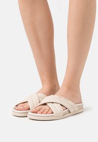 Sandales à brides croisées beiges avec une texture lisse et une semelle en couches dotée d'un accent blanc. Nom de la marque visible sur le côté.