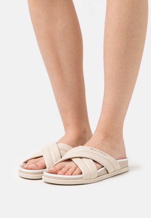 Sandales à brides croisées beiges avec une texture lisse et une semelle en couches dotée d'un accent blanc. Nom de la marque visible sur le côté.