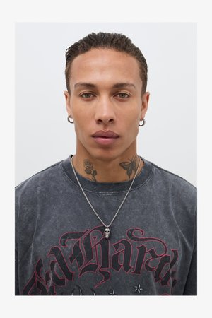 Halli suur T-särk, millel on punane graafiline trükk, hõbedane kettkaelakee, millel on kolju ripats, nähtavad tätoveeringud kaelal, sile nahk, lühikeste juustega soeng.