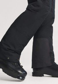 Čierne vodoodolné nohavice s priliehavým strihom, spevnenou dolnou časťou nohavíc a cvokmi. Spárované so silnými čiernymi horolezeckými topánkami.