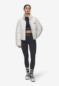 Hvit puffervest med høy krage, med glidelås i front og liten brystlomme, kombinert med svarte leggings og joggesko.