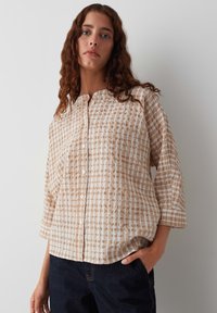 Chemise à carreaux beige et blanche avec un devant boutonné, des manches ¾ et une coupe ample. Tissu texturé avec un motif à grille subtil.