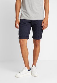 Uomo che indossa pantaloni corti a righe blu scuro, una maglietta grigia chiara e scarpe da ginnastica bianche, in piedi su un pavimento bianco contro un muro semplice.