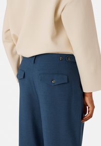 Blauwe op maat gemaakte broek met twee achterzakken, elk voorzien van klepjes en knopen. De stof lijkt glad en gestructureerd.