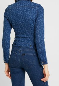 Dark denim jacket with a textured geometric pattern, featuring a collared neck and fitted design, paired with dark jeans and contrasting stitching.