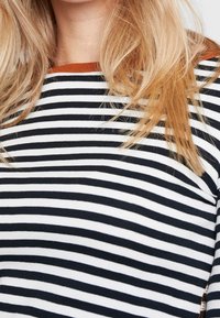 Maglietta a maniche lunghe a righe bianche e nere con scollo marrone. Il tessuto appare morbido e liscio, con un design aderente.