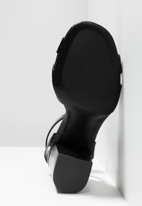 Sandale à talon haut noire avec une semelle texturée et un talon carré, positionnée droite contre un fond blanc dans un coin.
