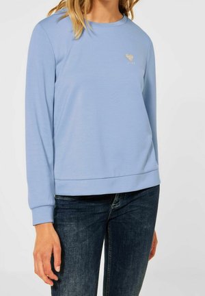 Kobieta w jasnoniebieskiej bluzie z długim rękawem z małym logo serca i strzały na piersi, w ciemnoniebieskich dżinsach.