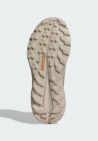 Tan spalvos guminis batų padas su giliomis, daugiakryptėmis raštinėmis, turinčiomis centrinę griovelį ir oranžine logotipo etikete. Lengvas, tekstūruotas paviršius.