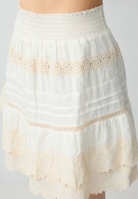 Jupe en coton blanc avec taille smockée, design superposé, bord en dentelle beige et accents floraux brodés à l'ourlet.