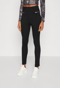 Zwarte high-waisted broek met een gestructureerde tailleband, twee ritszakken en een slim-fit ontwerp, gecombineerd met witte sneakers.