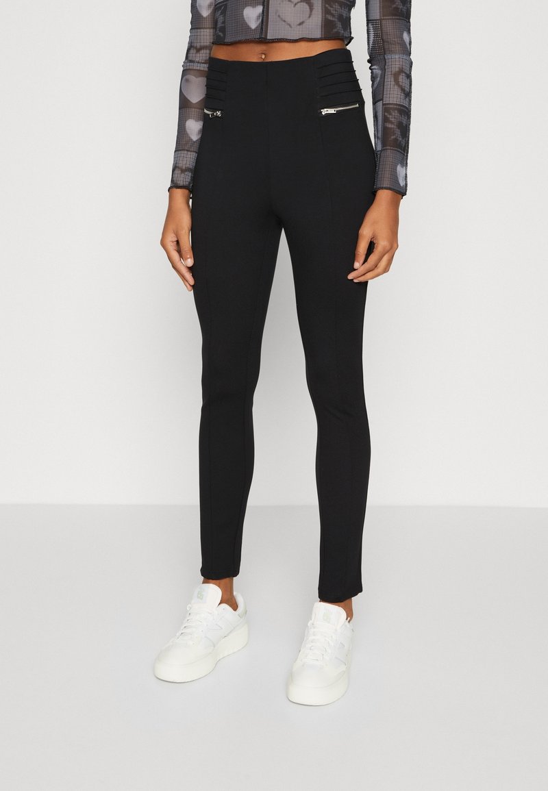 Zwarte high-waisted broek met een gestructureerde tailleband, twee ritszakken en een slim-fit ontwerp, gecombineerd met witte sneakers.