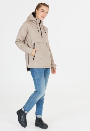 Ljusbeige hoodie-jacka med en känguruficka, justerbara dragsnören, ficka med dragkedja på bröstet, och en avslappnad passform. Kombinerad med blå jeans och svarta stövlar.