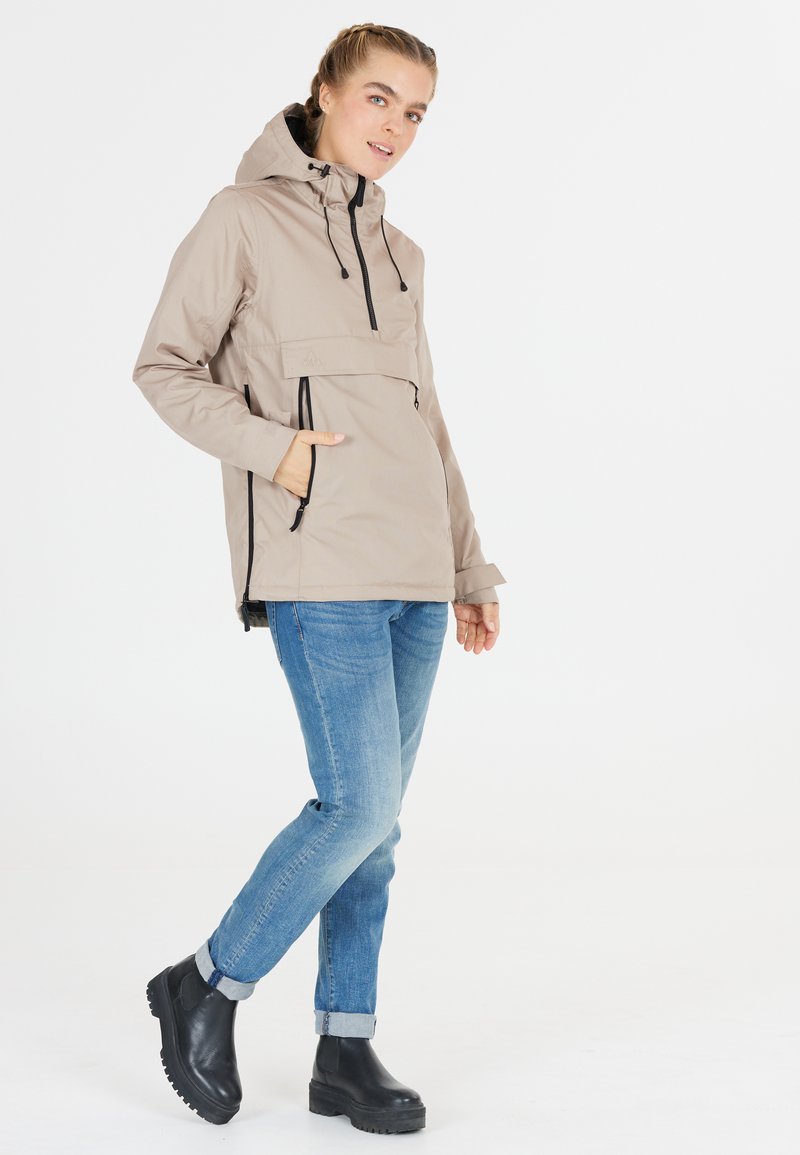 Lichtbeige hoodie met kangaroo pocket, verstelbare trekkoorden, een ritsvak op de borst en een ontspannen pasvorm. G gedragen met blauwe jeans en zwarte laarzen.