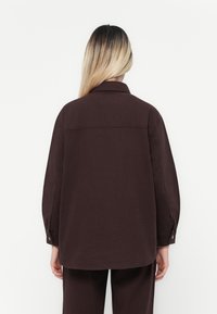 Mørkebrun skjorte med lange ermer, krage, knappelukking på ermene og rakskåret. Stoffet har en glatt tekstur og minimal detalj.