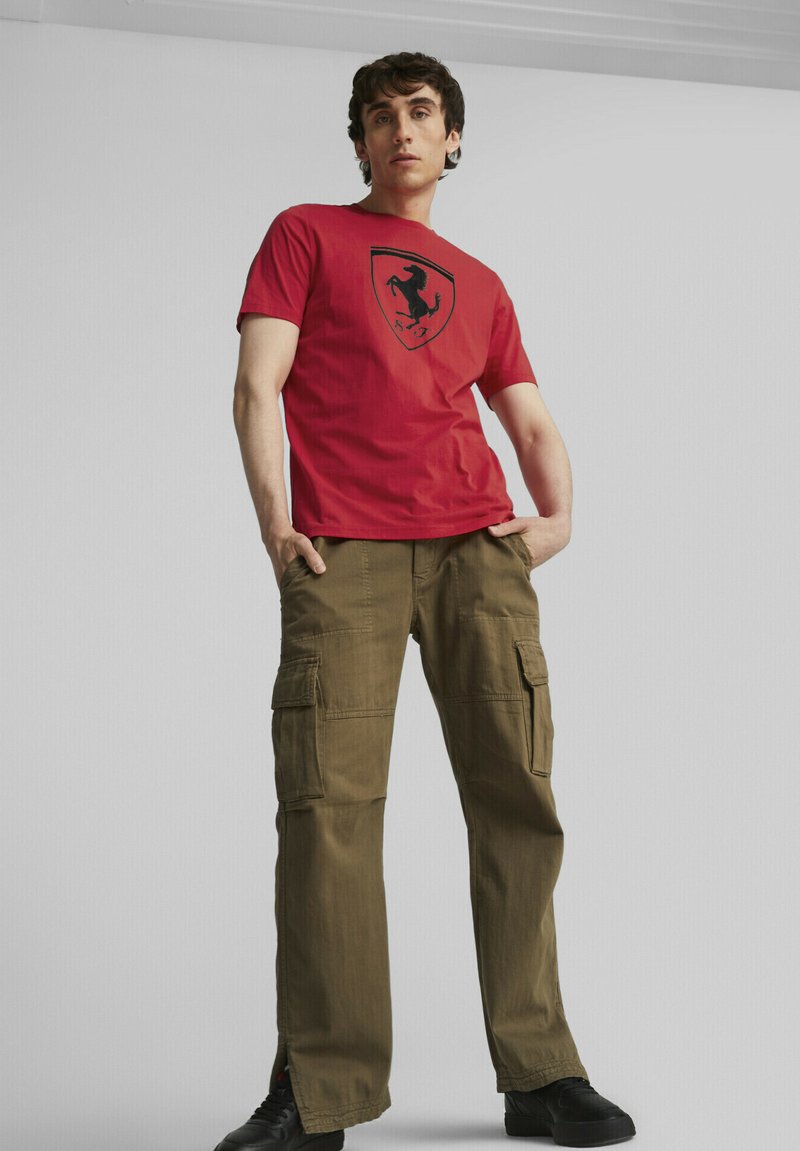 Czerwona bawełniana koszulka z czarnym logo Ferrari, połączona z oliwkowymi spodniami typu cargo z wieloma kieszeniami i czarnymi butami.