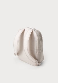 Vaalea beige reppu, jossa on teksturoitu pinta, vetoketjukiinnitys, pehmustetut hihnat ja sivutasku. Pyöreä muoto ja hillityt ompeluyksityiskohdat.