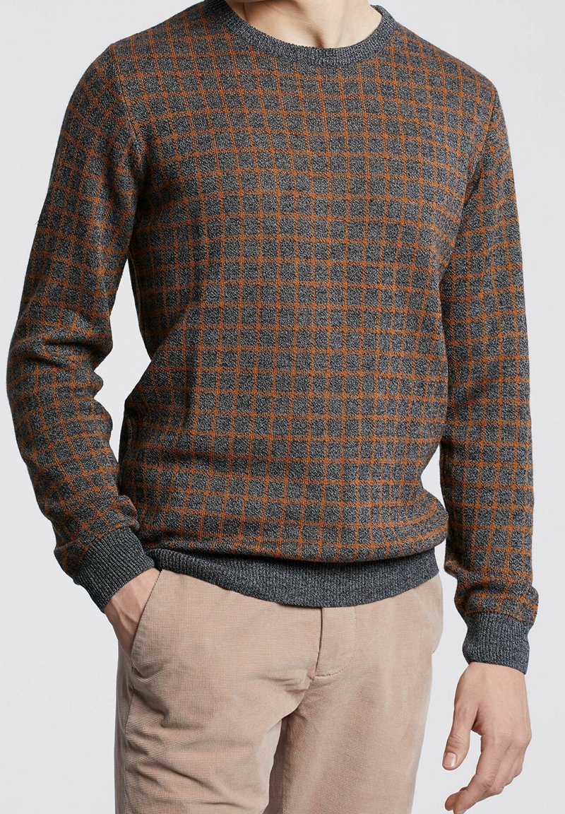 Homme portant un pull à carreaux gris et orange avec un pantalon beige, une main dans la poche, sur un fond clair uni.
