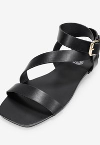 Sandal in pelle nera con cinturini incrociati e fibbia laterale. Suola piatta con texture liscia e design minimalista.