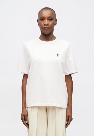 Person, der ein lockeres weißes T-Shirt mit einer kleinen gestickten Katze auf der Brust in einer blauen Jacke trägt, kombiniert mit hellen, gefalteten Hosen.
