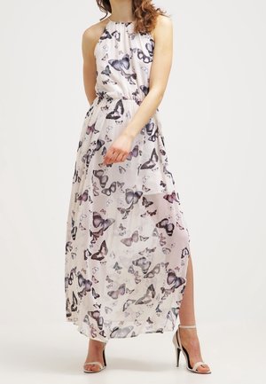 Vrouw in een mouwloze witte maxi-jurk met paarse en zwarte vlinderprint, zijsplit en witte sandalen met hoge hak.