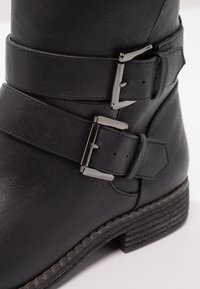 Botín de cuero negro con dos correas ajustables con hebillas metálicas, una suela texturizada y detalles de costura a lo largo de la base.
