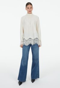 Cream blouse with high ruffled collar and lace hem. Long sleeves with lace accents. Paired with wide-leg denim jeans and pointed black shoes.
