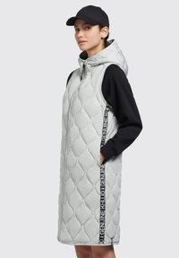 Helle graue gesteppte ärmellose Jacke mit Kapuze, ausgestattet mit einem schwarzen Seitenstreifen mit Text und seitlichen Taschen; kombiniert mit einem schwarzen Langarmoberteil.