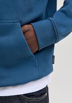 Μπλε φούτερ με κουκούλα, ριγέ μανίκια, πλαϊνές τσέπες και μια μικρή μαύρη ετικέτα. Το ύφασμα φαίνεται μαλακό και με υφή. Φορεμένο πάνω από ένα λευκό πουκάμισο.