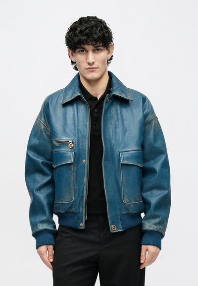 Νέος άνδρας με σπαστά μαλλιά που φοράει μπλε δερμάτινο μπουφάν πάνω από μαύρο πουκάμισο, στέκεται μπροστά σε απλό λευκό φόντο.